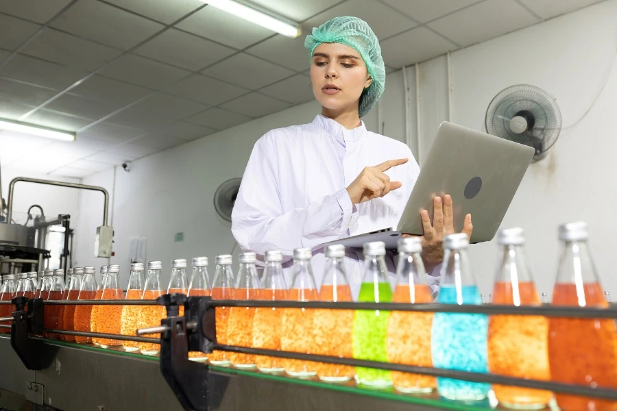 young woman factory worker looking basil seed drink and using laptop computer for checking quality in beverage factory